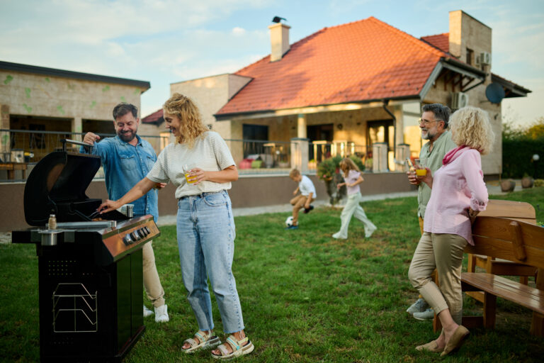 Family is enjoying a barbecue party in their backyard, with the parents grilling food and the children playing soccer. The warm evening light and lush green lawn create a cheerful atmosphere